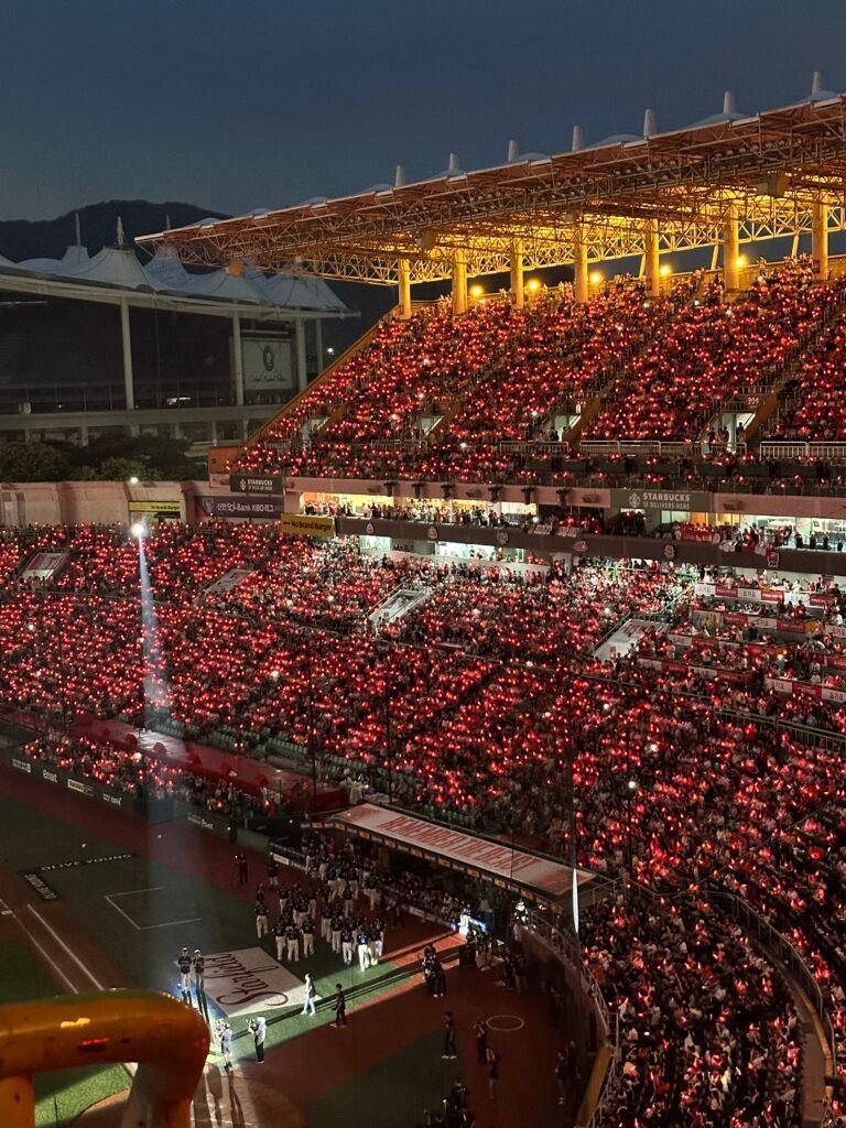 As the sun sets, a baseball player's retirement ceremony begins. Fans warmly welcome him, holding glowing red cheering sticks in the stands.