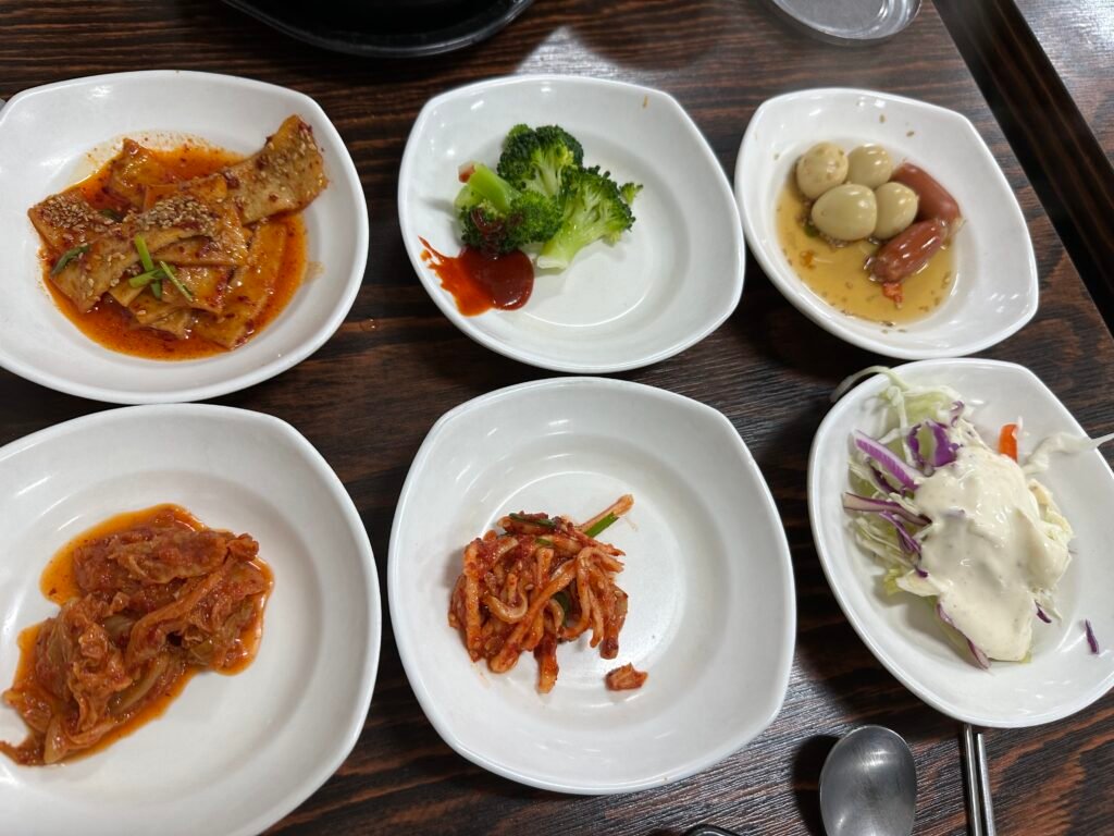 Six Korean side dishes including stir-fried fish cakes, broccoli, braised beef, stir-fried kimchi, seasoned radish, and salad