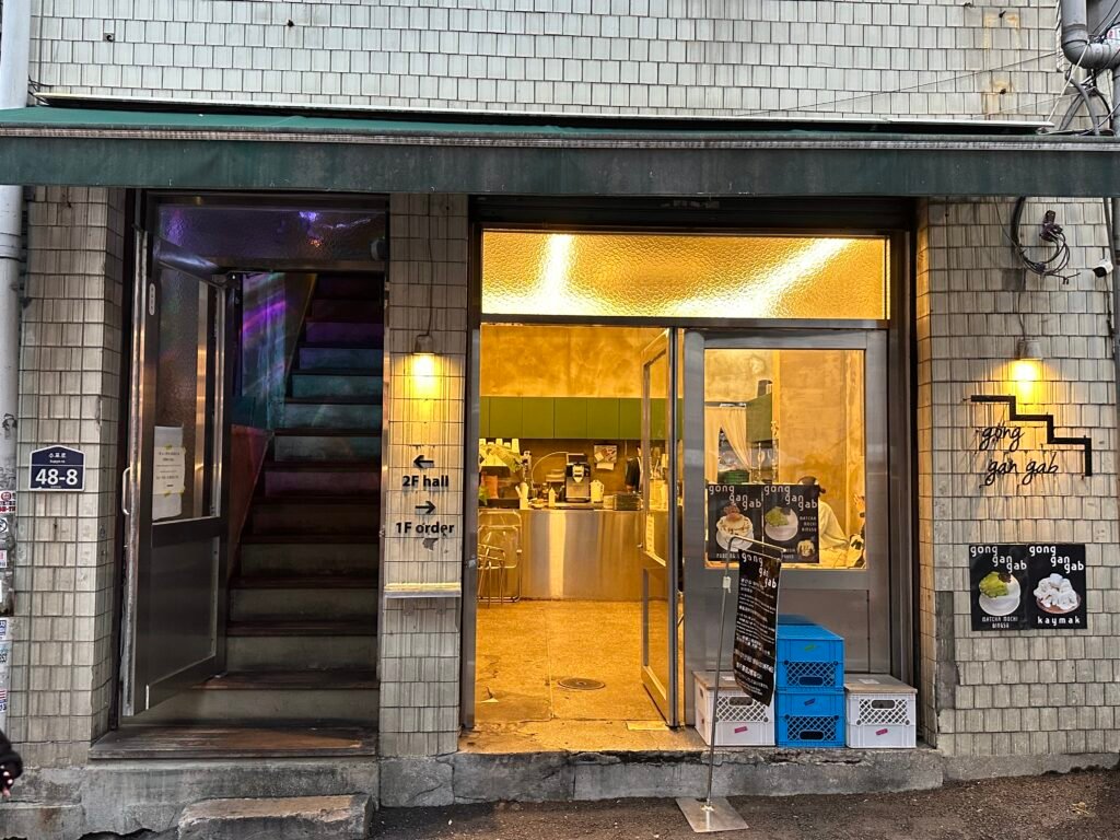 A cozy café glowing on the first floor along an Euljiro-style alley, with stairs leading up to the second-floor seating.