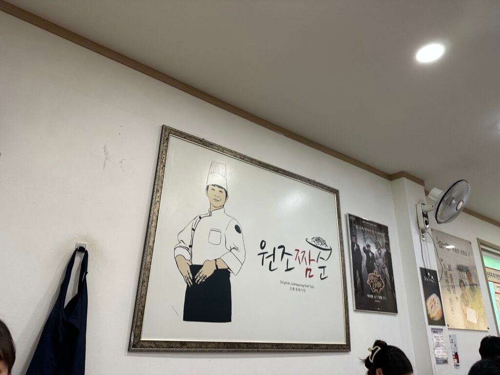 Framed sign reading “Wonjo Jjamsun,” meaning the original Jjamppong Sundubu, displayed on a white wall with TV show posters