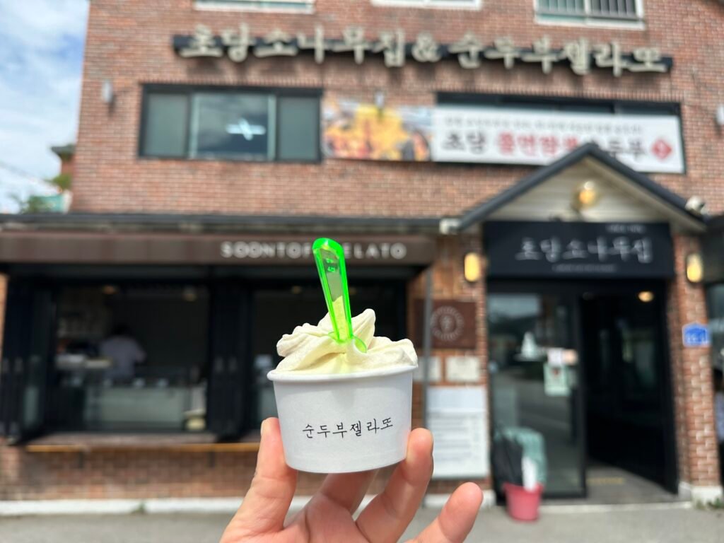 Hand holding a cup of sundubu gelato with Chodang Sonamujip shop in the background