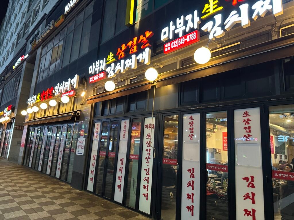 A restaurant exterior featuring a white and red Korean sign that reads “Mabuja Fresh Pork Belly Kimchi Stew,” with large open windows lining the first floor