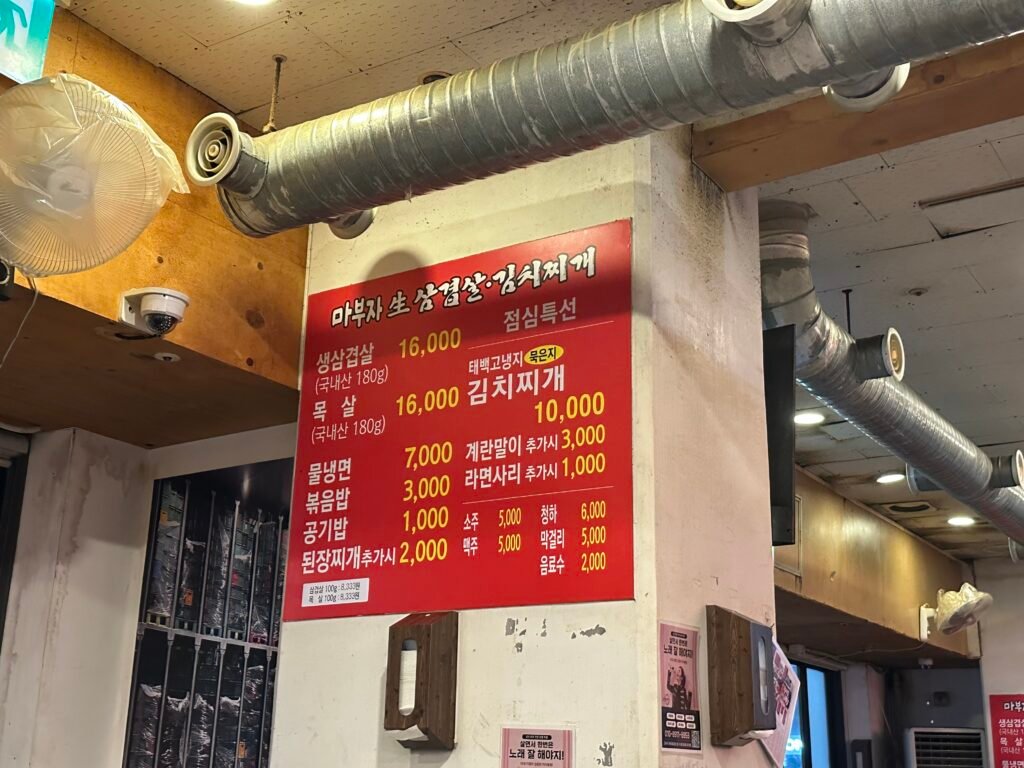 A bold red menu board on a white wall, clearly displaying items like pork belly priced at 16,000 won and soybean paste stew at 2,000 won